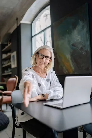 GPT
A smiling woman with shoulder-length, straight, light hair wearing glasses and a white blouse with black polka dots extends her arm, as if to greet someone with a handshake. She sits at a black table with a laptop open in front of her, in a well-lit, modern room with a large, abstract painting in dark tones on the wall behind her. The environment suggests a creative and professional setting.